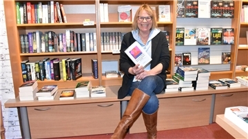 Eine blonde Frau sitzt vor einem Bücherregal und hat ein Buch in der Hand.
