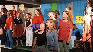 Kinder stehen vor einem blauen Bauwagen auf einer Theaterbühne. 