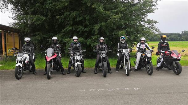Zu sehen sind sieben Motorradfahrer, die auf aufgereiht auf einem Parkplatz stehen. 