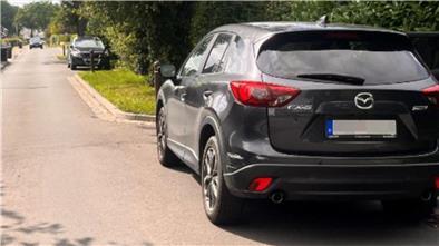 Ein schwarzes Auto steht rechts in einer Parkbucht und hat eine Schramme auf der linken Seite der hinteren Stoßstange.