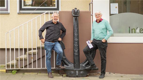 Zwei Männer, die vor einem Anker stehen. Der Anker steht vor einem gelben Gebäude.