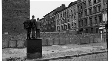 Drei Männer blicken über die Berliner Mauer gen Osten. Doch was genau sie dort sehen, bleibt verborgen. Sind sie nur neugierig oder nehmen sie Kontakt zu ihren Angehörigen hinter der Mauer auf?