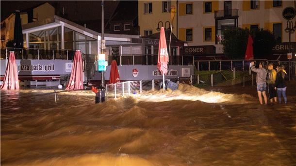 Viele Tote bei Unwettern - Häuser in der Eifel eingestürzt Drei Personen machen Fotos und beobachten die Wassermassen, die sich über eine Straße und ein Restaurant in Euskirchen ausbreiten.