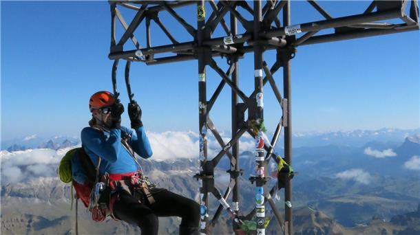 Ein Bergsteiger macht an seinen Eisgeräten einen Klimmzug am Gipfelkreuz der Marmolada.