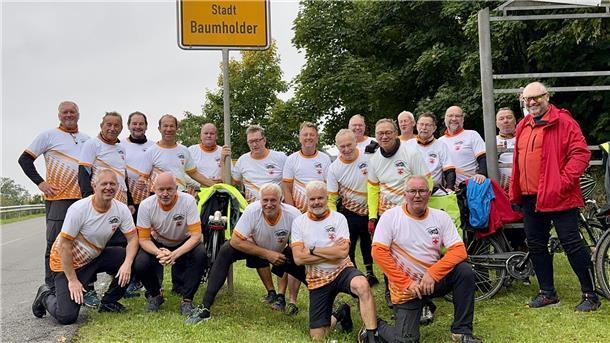 Eine Gruppe von 19 Radfahrern in einheitlichen Trikots steht lächelnd unter dem Ortsschild der Stadt Baumholder. Im Hintergrund sind Bäume und mehrere Fahrräder zu sehen.