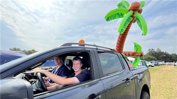 Zwei Männer in einem Auto. Auf der Ladefläche sind zwei Palmen aus Plastik.