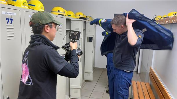  In einer Umkleide eines Hilfswerks zieht sich ein junger Mann eine blaue Einsatzjacke über, während ein Kameramann mit Kappe und schwarzem T-Shirt ihn mit einer professionellen Kamera filmt. Im Hintergrund sind Spinde mit gelben Schutzhelmen zu sehen.