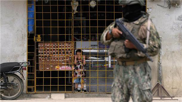 Vor Wahl: Weiterer Lokalpolitiker in Ecuador getötet Ein Junge schaut hinter den Gitterstäben eines geöffneten Elektronikgeschäfts hervor, während ein Soldat an einem von der Armee eingerichteten Sicherheitskontrollpunkt Wache steht.