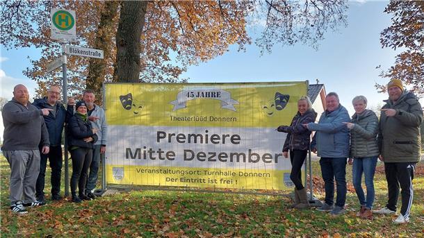 Eine Gruppe von Menschen steht in einer herbstlichen Parklandschaft vor einem großen gelben Banner. Auf dem Banner steht in schwarzer Schrift: „Premiere Mitte Dezember“. Die Menschen lächeln und zeigen auf das Banner, um die Veranstaltung anzukündigen.