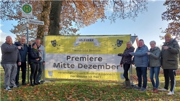 Eine Gruppe von Menschen steht in einer herbstlichen Parklandschaft vor einem großen gelben Banner. Auf dem Banner steht in schwarzer Schrift: „Premiere Mitte Dezember“. Die Menschen lächeln und zeigen auf das Banner, um die Veranstaltung anzukündigen.