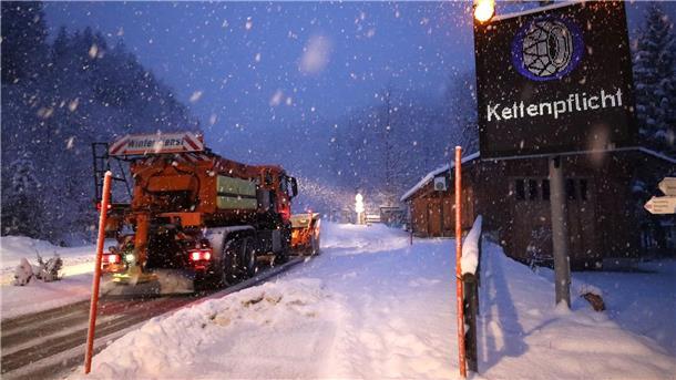 Schneefall in Bayern - erhebliche Lawinengefahr in den Alpen Ein Schild weist auf die Schneekettenpflicht am Riedbergpass im bayerischen Landkreis Oberallgäu hin, während daneben ein Räumfahrzeug den Schnee von der Straße räumt.