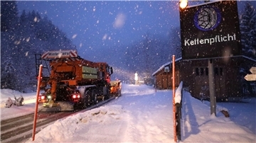 Ein Schild weist auf die Schneekettenpflicht am Riedbergpass im bayerischen Landkreis Oberallgäu hin, während daneben ein Räumfahrzeug den Schnee von der Straße räumt.