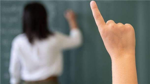 Ein Schüler meldet sich, während die Lehrerin an die Tafel schreibt.