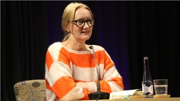 Eine Frau mit Brille und blondem Haar sitzt an einem Tisch mit Mikrofon. Sie trägt einen Pullover mit orange-weißen Streifen, vor ihr stehen ein Glas Wasser und eine Flasche.