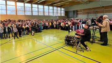 Eine große Gruppe von Kindern und einige Erwachsene versammeln sich in einer Sporthalle. Im Vordergrund ist ein Schüler an einem Schlagzeug zu sehen, begleitet von weiteren Musikern mit Gitarren und einem Keyboard. 