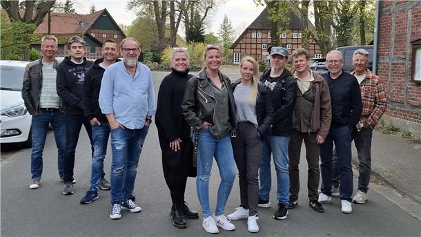 Gruppe von elf Personen, darunter Männer und Frauen, steht lächelnd auf einer Dorfstraße vor Fachwerkhäusern; sie tragen Freizeitkleidung und wirken gut gelaunt.