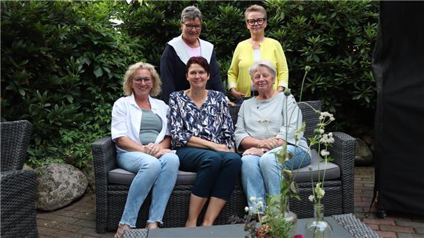 Drei Frauen sitzen nebeneinander auf einer Bank und zwei Frauen stehen dahinter.