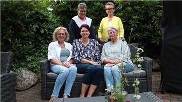 Drei Frauen sitzen nebeneinander auf einer Bank und zwei Frauen stehen dahinter.