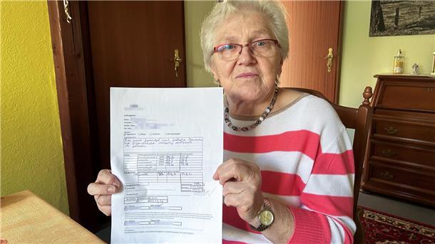 Eine ältere Frau blickt in die Kamera und zeigt die teure Rechnung.