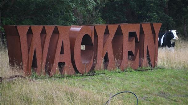 Ein Wacken-Schriftzug aus Metall steht neben einer Plastik-Kuh auf dem Festivalgelände.
