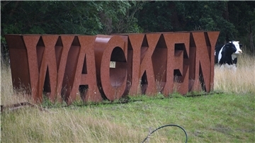 Ein Wacken-Schriftzug aus Metall steht neben einer Plastik-Kuh auf dem Festivalgelände.