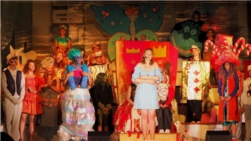 Schülerinnen und Schüler des Gymnasiums Wesermünde in bunten Kostümen auf der Bühne während des Musicals „Alice“. Im Mittelpunkt steht eine Schülerin im blau-weißen Kleid als Alice, umgeben von fantasievoll verkleideten Figuren wie Karten, Tieren und Fabelwesen vor einer farbenfrohen Kulisse.