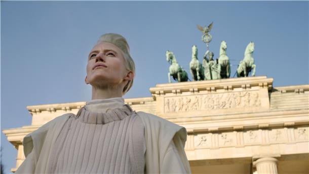 Eine Erlöserfigur im weißen Mantel mit einem Patronengurt um den Bauch - die Künstlerin selbst - schreitet neben einem gescheckten Esel durchs Brandenburger Tor - eine Szene aus Yael Bartanas Filminstallation „Malka Germania“.