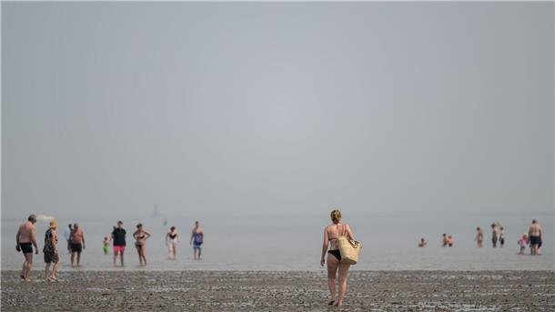Wissenschaftler erforschen Munition in Ost- und Nordsee Eine Frau geht bei sommerlichen Temperaturen am Nordseestrand von Schillig im Watt auf andere Strandgäste zu, die teilweise im Wasser sind.
