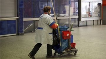Eine Gebäudereinigerin schiebt einen Wagen mit Reinigungsutensilien vor sich her.
