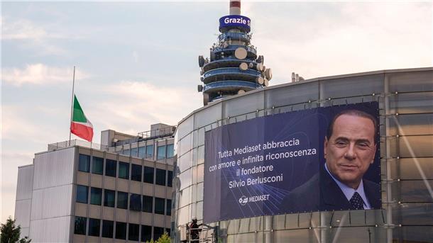 Nach Tod von Berlusconi: Italiens Regierung gefordert Eine italienische Flagge auf Halbmast weht vor einer Nachricht mit der Aufschrift „Grazie Silvio“ auf einem Turm des Fernsehsenders Mediaset neben einem großen Plakat.
