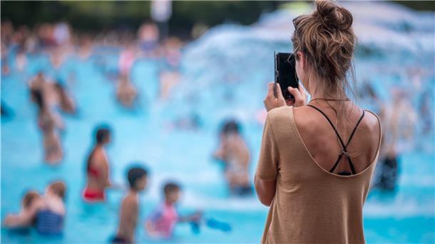 Eine junge Frau fotografiert mit ihrem Handy in einem Freibad. Hunderttausende Kinder und Erwachsene folgen minderjährigen Influencern auf Youtube, Instagram oder TikTok durch ihren Alltag, in den Park oder ins Schwimmbad. Hinter der Kamera, den Accounts und den Verträgen mit Unternehmen, die in den Videos ihre Produkte platzieren, stehen oft die Eltern.