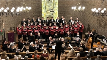 Ein Chor aus Männern und Jungen in schwarzer und roter Chorkleidung steht in einer Kirche vor einer Wand mit einem großen Mosaik. Vor dem Chor sitzt ein Orchester mit Musikern, die verschiedene Instrumente spielen, darunter Geigen, Cello und Kontrabass. Ein Dirigent steht in der Mitte und leitet das Ensemble. Im Vordergrund sind die Rücken von Zuschauern zu sehen, die das Konzert verfolgen.