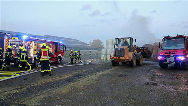 Ein Radlader transportiert einen Heuballen, während Feuerwehrleute neben ihrem Einsatzfahrzeug stehen. 