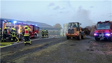 Ein Radlader transportiert einen Heuballen, während Feuerwehrleute neben ihrem Einsatzfahrzeug stehen. 