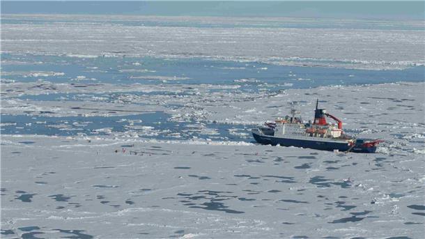Der Forschungseisbrecher „Polarstern“ liegt inmitten von arktischem Meereis, umgeben von Schollen und offenen Wasserflächen. In der Nähe arbeiten Personen auf dem Eis an Messstationen.
