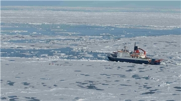 Der Forschungseisbrecher „Polarstern“ liegt inmitten von arktischem Meereis, umgeben von Schollen und offenen Wasserflächen. In der Nähe arbeiten Personen auf dem Eis an Messstationen.