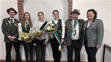 Sechs Mitglieder eines Schützenvereins stehen nebeneinander vor einer weißen Wand mit Ziegelbögen. Sie tragen traditionelle Schützenuniformen mit Orden und Schärpen. Drei Frauen in der Mitte halten Blumensträuße. 