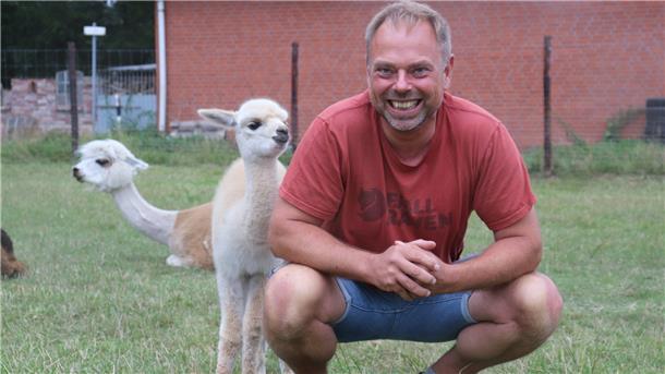 Ein Mann hockt auf einer Weide, hinter ihm zwei Alpakas.