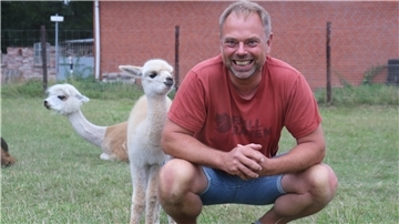 Ein Mann hockt auf einer Weide, hinter ihm zwei Alpakas.