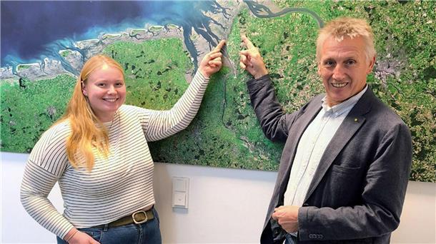 Eine junge Frau und ein älterer Mann stehen vor einer Satellitenkarte der Nordseeküste und zeigen lächelnd auf einen Punkt in der Region Wurster Nordseeküste.