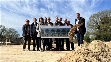 Menschen stehen auf einem Sandfeld, drei von ihnen halten ein Plakat. Der Bürgermeister schleudert Sand von einem Spaten.