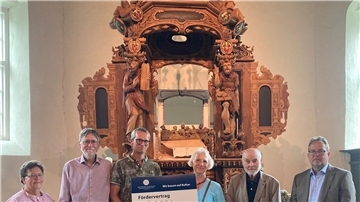 Sechs Frauen und Männer vor einem alten Altar mit symbolischen Scheck in den Händen
