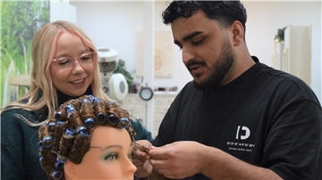 Ein Mann dreht die Haare einer Friseurpuppe mit Lockenwicklern auf. Eine Frau schaut ihm dabei zu. 