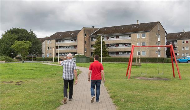 Zwei Männer mit kariertem Hemd bzw. rotem Polo-Hemd sind von hinten zu sehen. Sie durchschreiten einen Park mit roter Schaukel. Vor ihnen wartet ein Häuserblock.