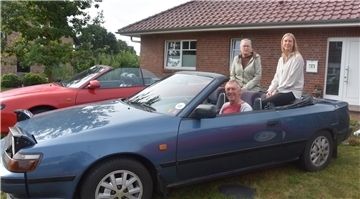 Ein Mann sitzt am Steuer eines Cabriolets, hinten auf dem Verdeck sitzen zwei Frauen.