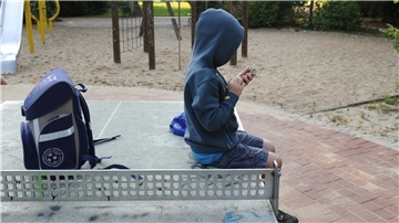 Ein Kind sitzt auf einer Tischtennisplatte und spielt mit dem Mobiltelefon. Daneben liegt der Schulranzen.