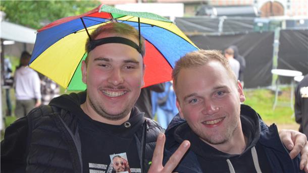 Zwei junge Männer lächeln in die Kamera. Der linke Mann hat einen bunten Regenschirm auf dem Kopf.