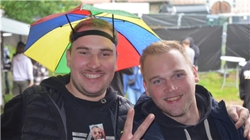 Zwei junge Männer lächeln in die Kamera. Der linke Mann hat einen bunten Regenschirm auf dem Kopf.