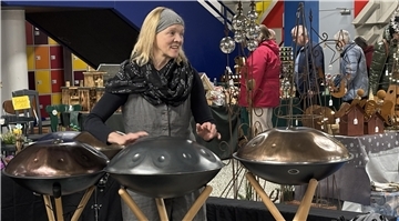Eine Frau spielt auf mehreren Handpans, die auf Ständern nebeneinander aufgereiht sind. Sie trägt ein Stirnband und einen Schal, während sie das Instrument in einer Ausstellungsumgebung präsentiert.