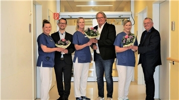 Drei in Anzügen gekleidete Männer übergeben in einem Krankenhausflur Blumensträuße an drei Pflegerinnen